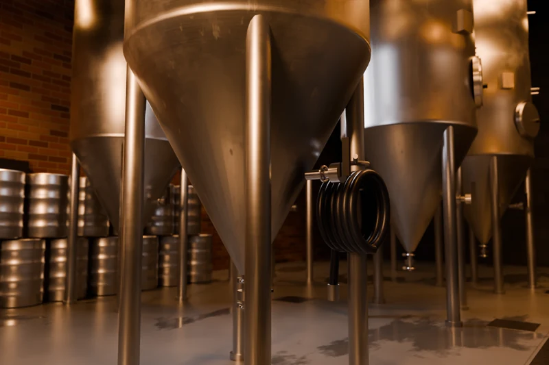 A close view of the brewhouse: stainless fermenting tanks on legs, stacked kegs, and a coil of dark hose hung on a tank leg.